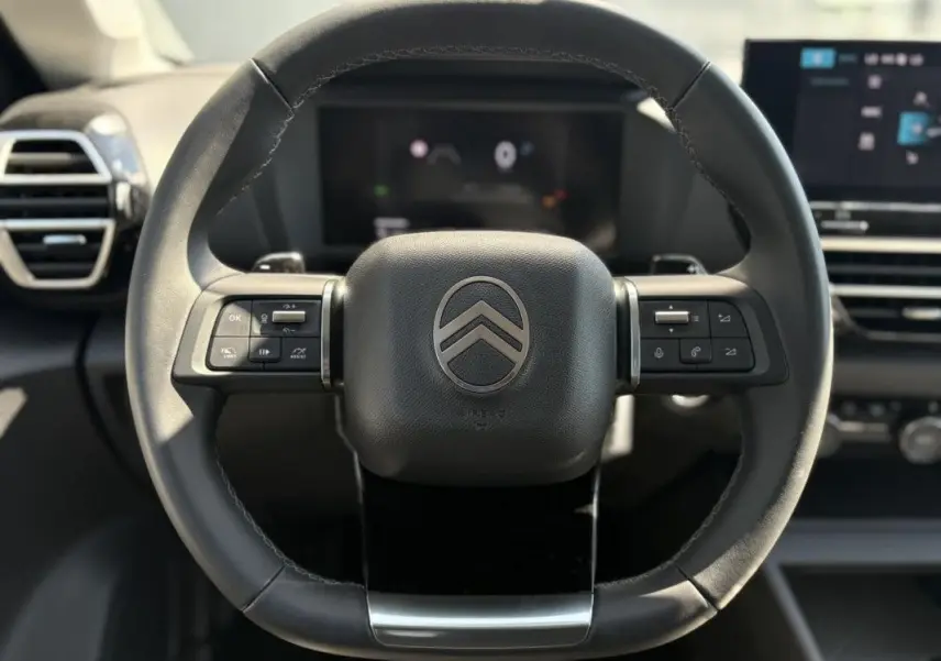 Gros plan sur le volant noir du Citroën C4 Hybrid 145 ch, avec commandes intégrées et tableau de bord numérique en arrière-plan.