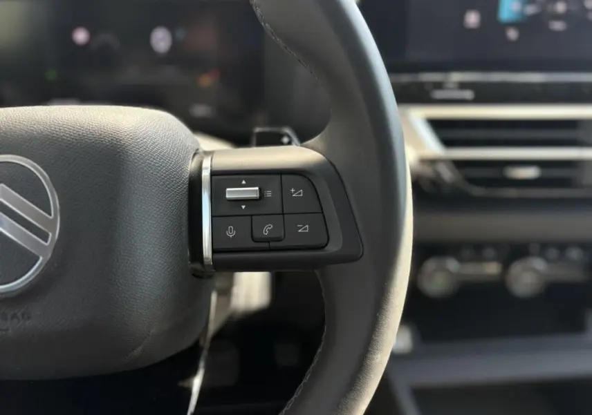 Gros plan sur les commandes gauche du volant de la Citroën C4 Hybrid 145 CH 2025, intérieur noir avec écran tactile en arrière-plan.