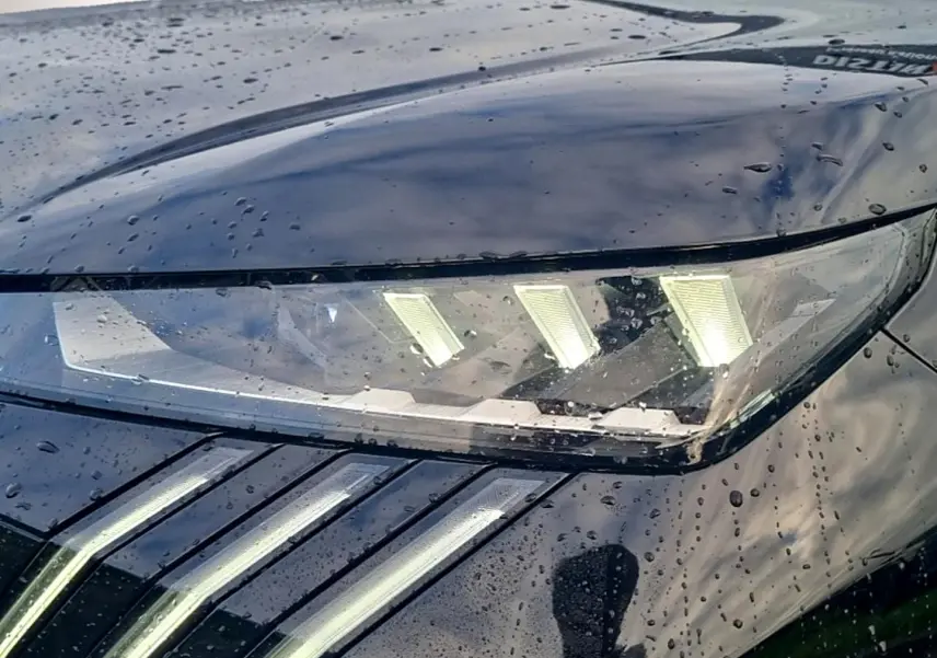 Gros plan sur le phare avant droit du Peugeot 5008 noir, avec ses trois griffes LED distinctives sous la pluie.