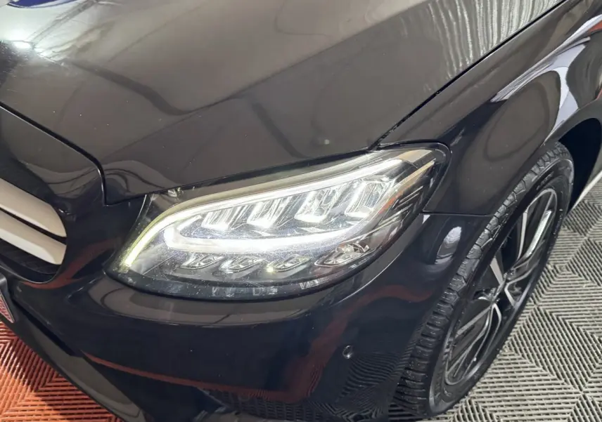 Gros plan sur le phare avant droit allumé d'une Mercedes Classe C Break noire, avec jante alliage visible.