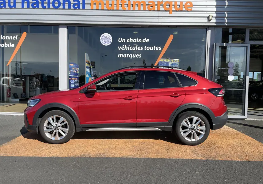 Vue de profil côté gauche d’un Volkswagen Taigo rouge Roi 2024 garé devant un showroom multimarque.