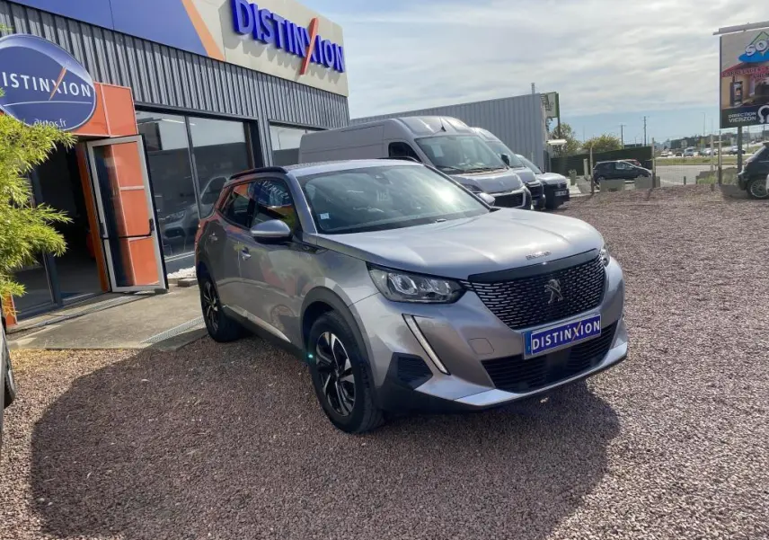 Peugeot 2008 gris clair vue 3/4 avant droit, avec calandre noire et jantes alliage noires, devant un bâtiment Distinxion.