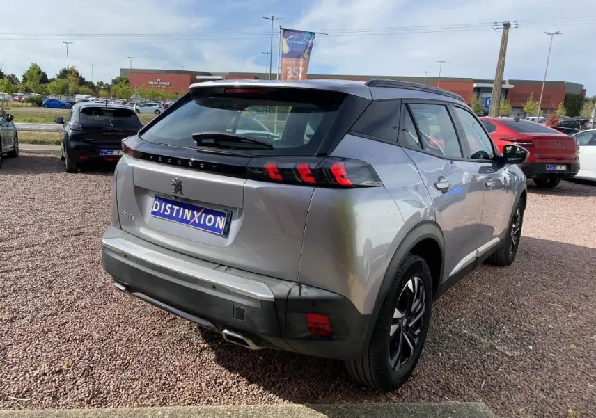 Vue 3/4 arrière droite d'un Peugeot 2008 gris clair avec barres de toit noires et feux arrière LED distinctifs.