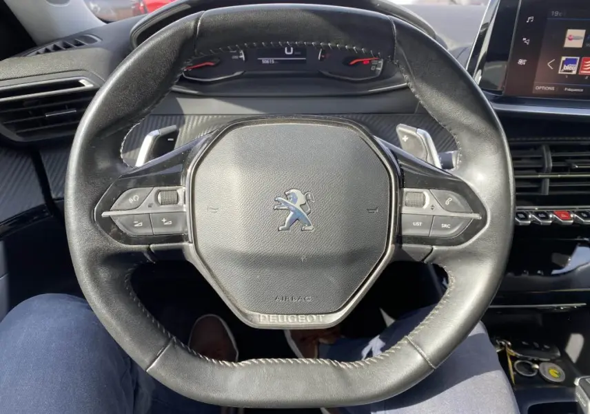 Vue rapprochée du volant cuir noir du Peugeot 2008 2020 avec commandes intégrées et tableau de bord numérique en arrière-plan.