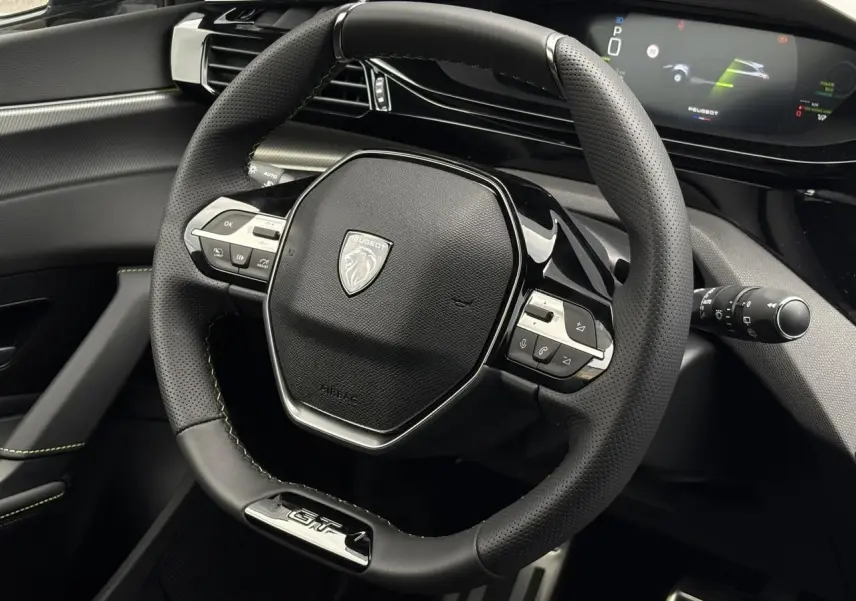 Vue rapprochée du volant GT noir avec surpiqûres vert Adamite et tableau de bord numérique du Peugeot 308 SW Hybrid 2026.