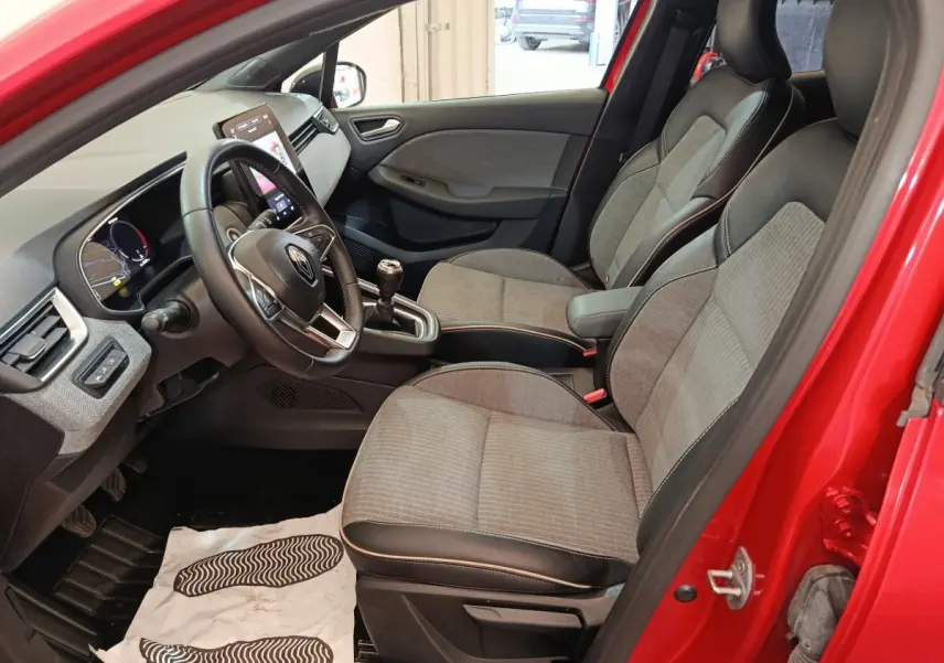 Vue intérieure côté conducteur d'une Renault Clio rouge 2024, sièges tissu gris avec accoudoir central et volant multifonctions.