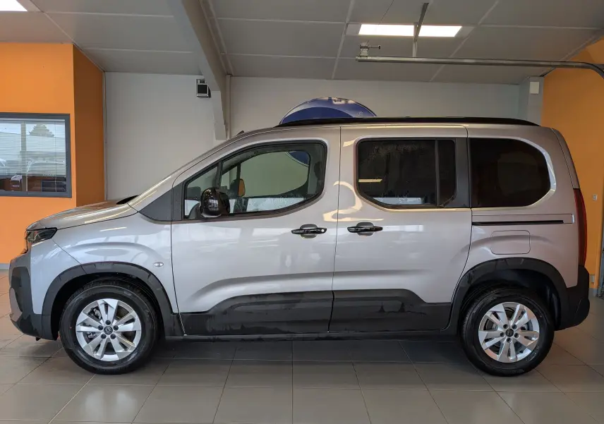 Vue latérale droite du Peugeot Rifter gris Artense 2025 avec barres de toit noires brillantes en intérieur showroom.