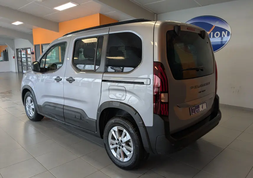 Vue 3/4 arrière droit du Peugeot Rifter gris Artense avec barres de toit noir brillant en intérieur showroom.