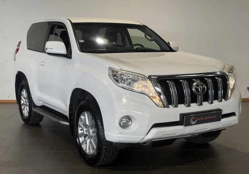 Toyota Land Cruiser blanc vue 3/4 avant droit, avec calandre chromée et phares allumés, intérieur visible.