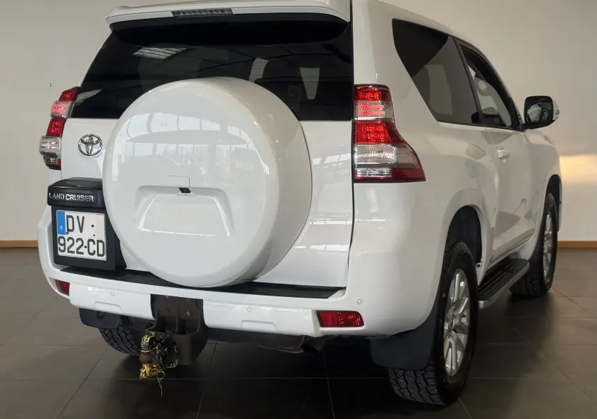Toyota Land Cruiser blanc vu 3/4 arrière droit, avec roue de secours intégrée à la porte et attelage visible.