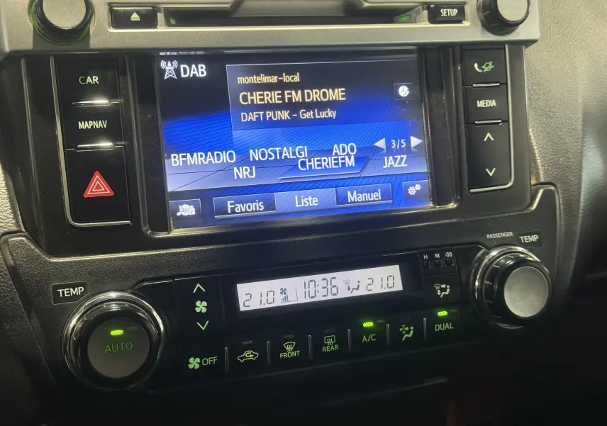 Gros plan sur l’écran multimédia et la console de climatisation du Toyota Land Cruiser blanc, affichant une radio DAB et la température à 21°C.