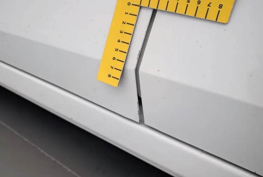 Détail de la portière blanche de la Dacia Sandero 2022 avec un écart anormal et un outil de mesure jaune posé dessus.