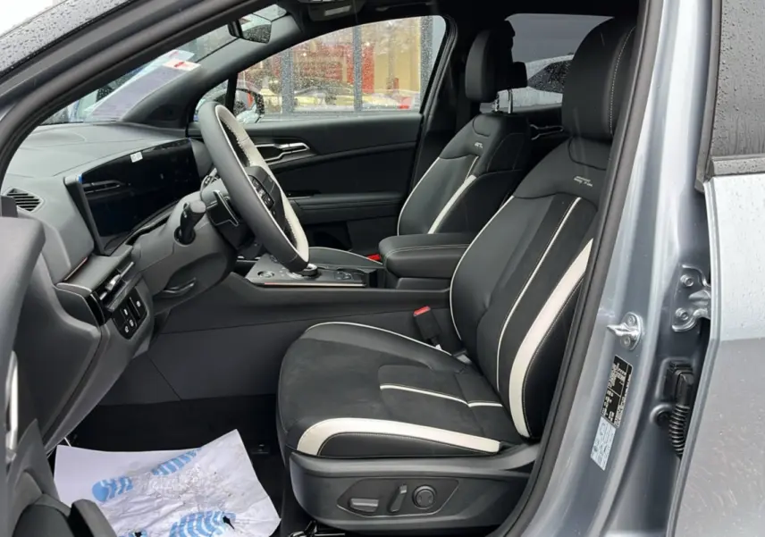 Vue intérieure côté conducteur de la KIA Sportage 2026 gris perle, sièges sport noirs avec surpiqûres blanches et volant cuir multifonctions.