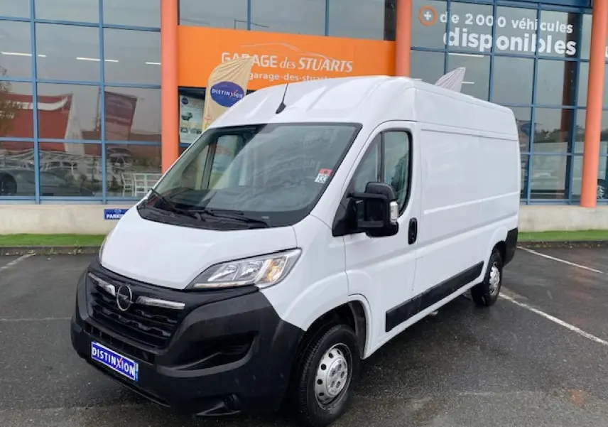 Vue 3/4 avant droit d’un Opel Movano Fourgon blanc avec porte latérale coulissante et pare-chocs noir, stationné devant un garage.
