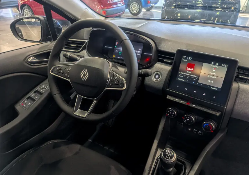 Vue intérieure côté conducteur de la Renault Clio V 2025, avec volant multifonction, écran tactile et levier de vitesse manuel.