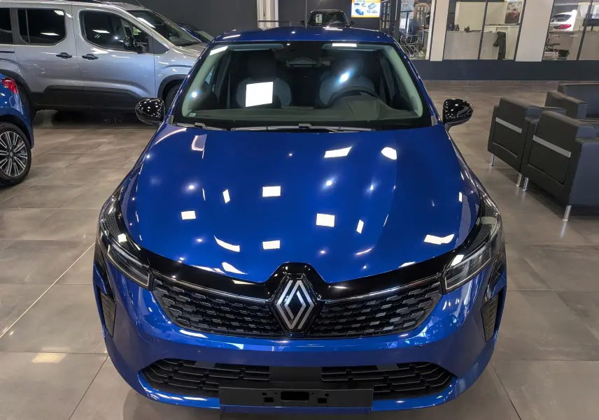 Vue avant d'une Renault Clio V Bleu Iron 2025 en showroom, avec calandre noire et logo Renault central brillant.