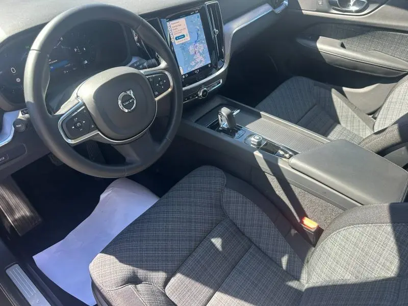 Vue intérieure côté conducteur de la Volvo S60 B4 2024 avec sièges tissu gris à motif, volant multifonction et écran tactile central.