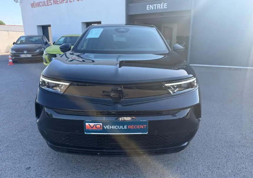 Vue avant d’un Opel Mokka électrique noir 2025, avec calandre lisse et phares LED fins, stationné devant un garage.