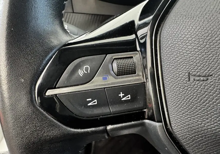 Gros plan sur les commandes gauche du volant cuir noir du Peugeot 2008 blanc, avec boutons de volume et reconnaissance vocale.