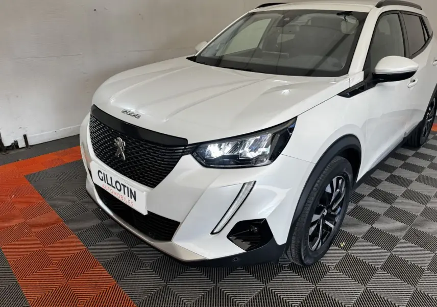 Peugeot 2008 blanc en 3/4 avant droit, avec calandre noire et phares LED allumés sur sol à damiers.