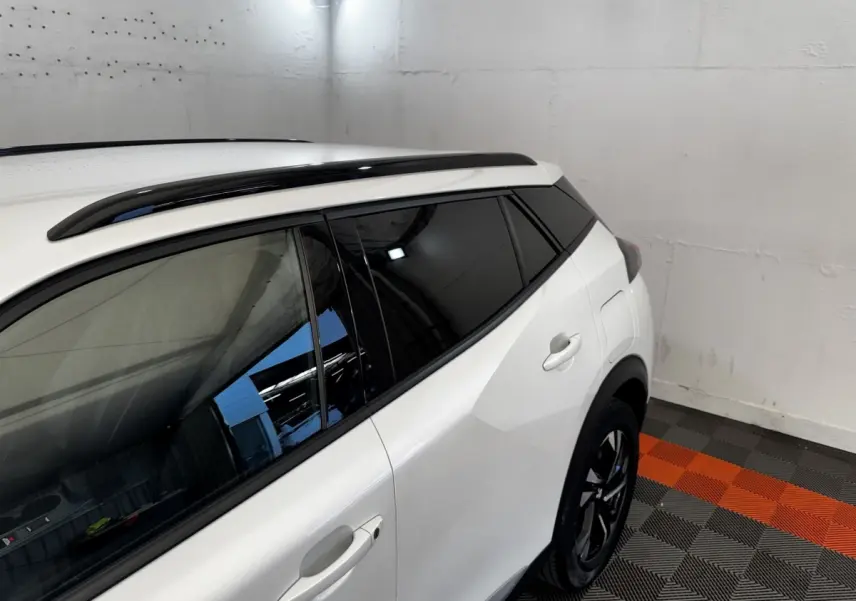 Vue 3/4 arrière droite d’un Peugeot 2008 blanc avec vitres teintées et barres de toit noires dans un garage.