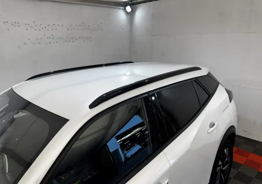 Vue 3/4 arrière droite du Peugeot 2008 blanc avec barres de toit noires et vitres teintées dans un garage.