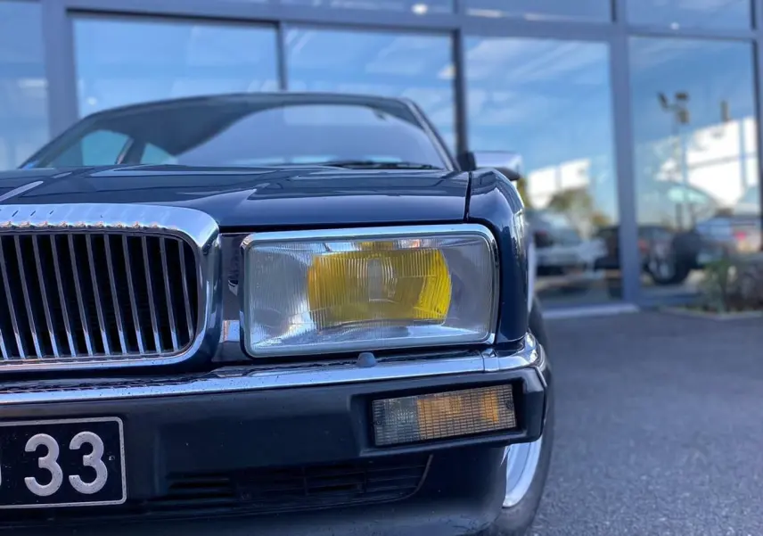 Gros plan sur l'avant droit bleu d'une Jaguar XJ6 3.6 Sovereign 1987, mettant en valeur le phare jaune et la calandre chromée.