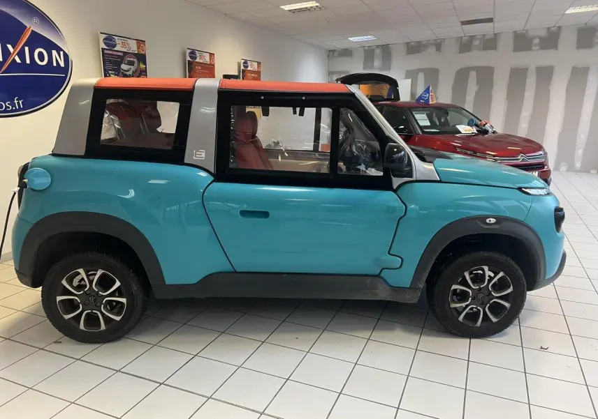 Profil côté gauche d'une Citroën E-MEHARI cabriolet bleu clair avec toit en toile rouge en concession.