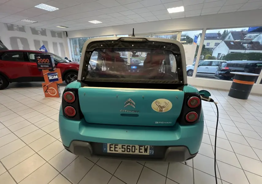 Vue arrière d'une Citroën E-MEHARI cabriolet bleu clair en recharge, avec toit en toile et autocollant cerf sur le hayon.