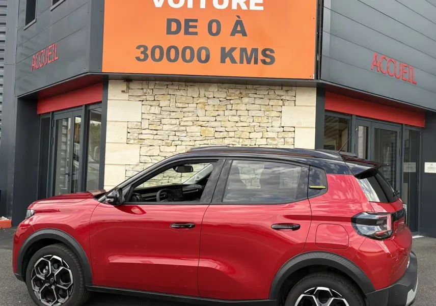 Vue de profil droit d'une Citroën C3 Turbo 100 BVM6 MAX rouge avec toit noir et jantes distinctives, devant un bâtiment.