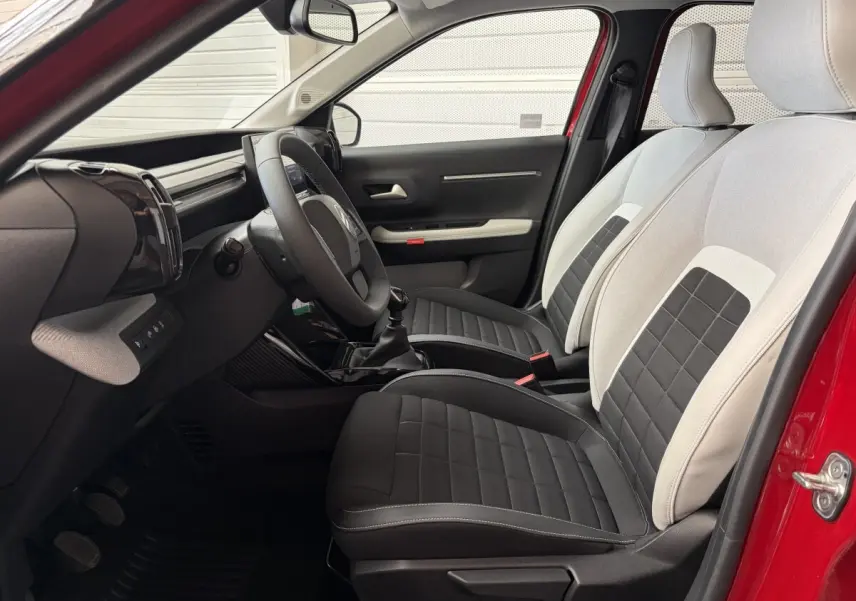 Vue intérieure côté conducteur d'une Citroën C3 rouge 2025, sièges bi-ton noir et beige, volant cuir et tableau de bord moderne.
