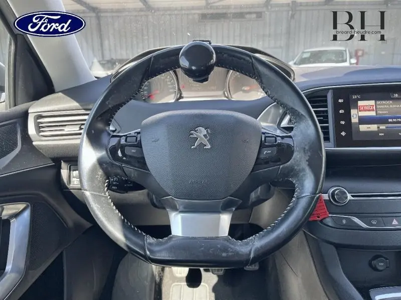 Vue intérieure centrée sur le volant cuir Peugeot avec commandes intégrées et écran tactile multifonction en arrière-plan.
