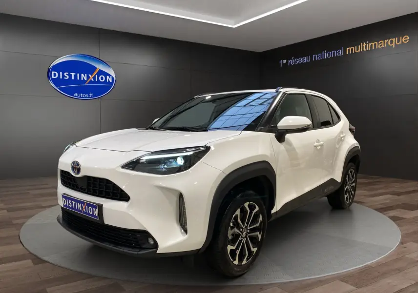 Toyota Yaris Cross blanc vue 3/4 avant droit, avec jantes alu 17 pouces et phares LED allumés dans showroom.