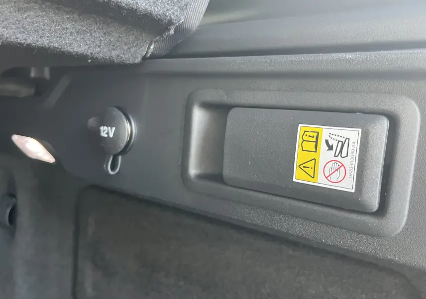 Détail intérieur du coffre du Jaguar F-PACE blanc 2018, prise 12V et commande avec avertissement de sécurité.