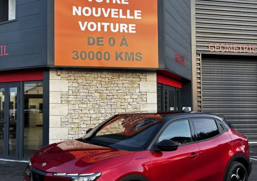 Vue 3/4 avant droite d'une Alfa Romeo Junior rouge avec toit noir, jantes noires et calandre distinctive devant un bâtiment commercial.