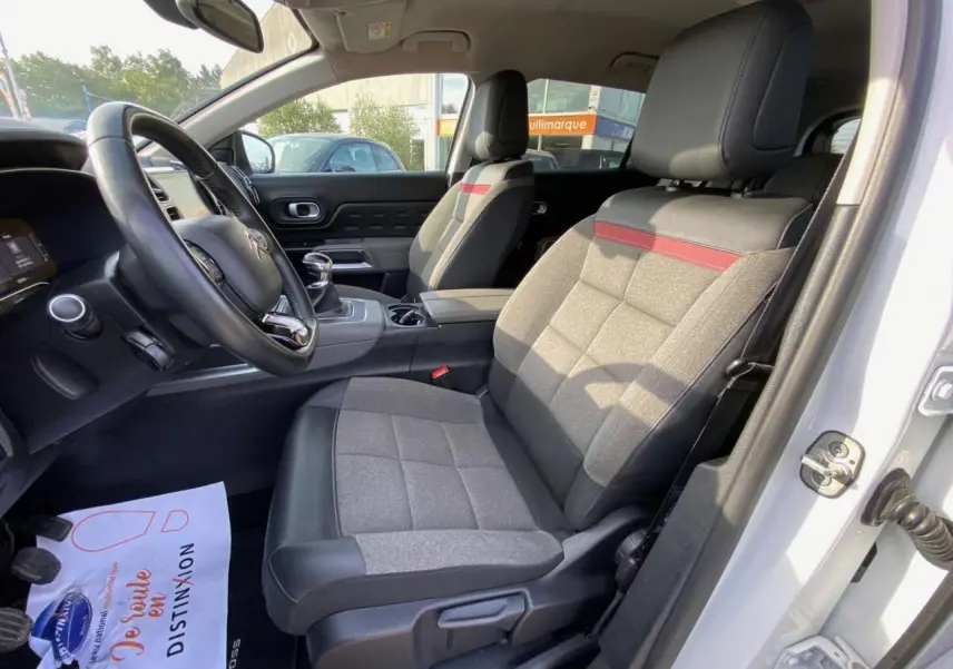 Intérieur avant droit du Citroën C5 Aircross blanc, sièges tissu et cuir gris avec bande rouge, volant et levier manuel visibles.