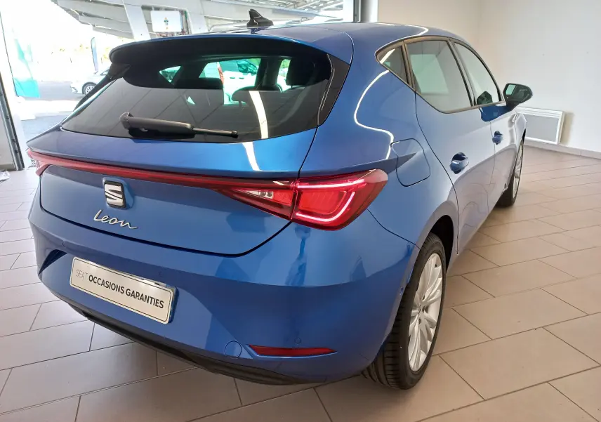 Vue 3/4 arrière droite d'une SEAT Leon 2025 bleu saphir avec feux arrière allumés et vitres surteintées en intérieur showroom.