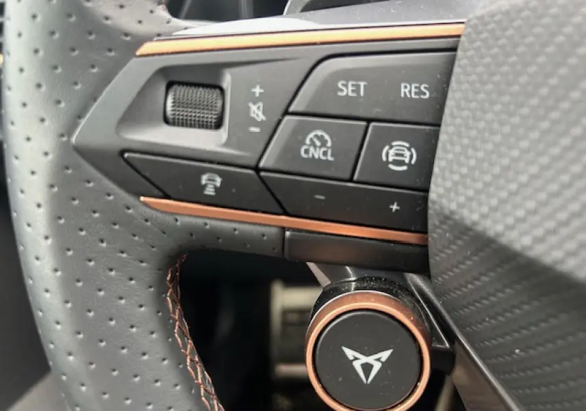 Gros plan sur le volant en cuir perforé gris avec commandes multifonctions et logo CUPRA cuivré au centre.