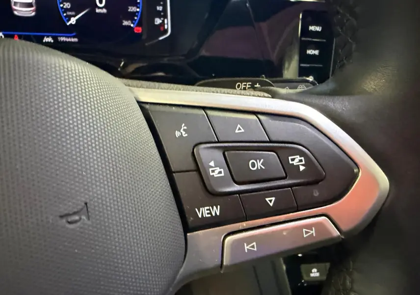 Gros plan sur les commandes multifonctions du volant cuir d’un Volkswagen Touran gris, tableau de bord numérique en arrière-plan.