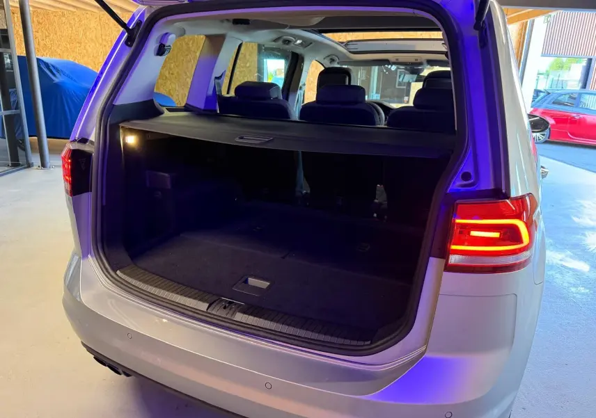 Vue arrière ouverte du coffre d'un Volkswagen Touran gris 2025 avec feux arrière LED allumés et toit ouvrant panoramique visible.