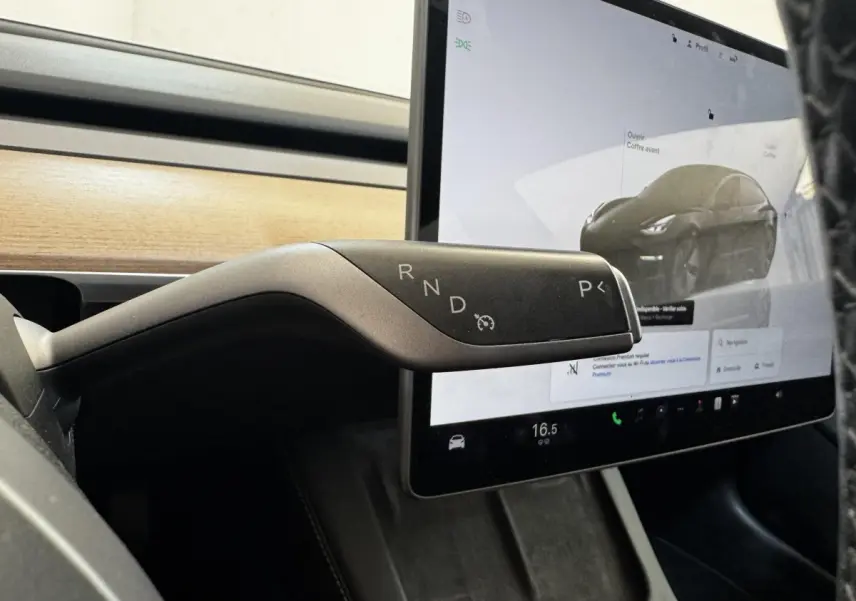 Gros plan sur la commande de boîte automatique noire de la Tesla Model 3 2023, avec écran tactile et tableau de bord bois en arrière-plan.