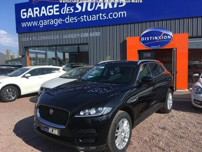 Jaguar F-PACE noir en 3/4 avant droit, jantes argentées, stationné devant un garage avec enseigne bleue et orange.