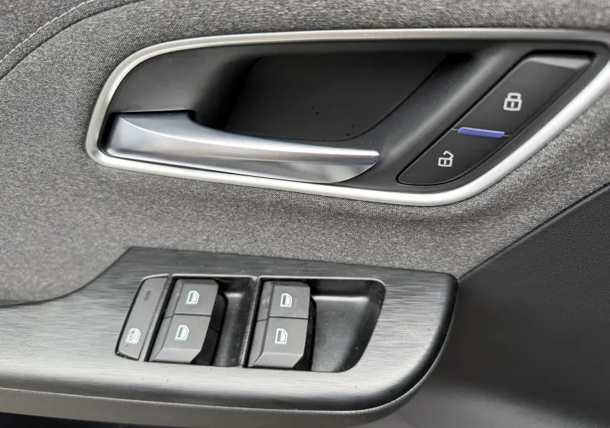 Poignée de porte intérieure et commandes des vitres en tissu gris sur MG ZS 1.5L Hybrid+ 2026, détails en plastique noir et argenté.