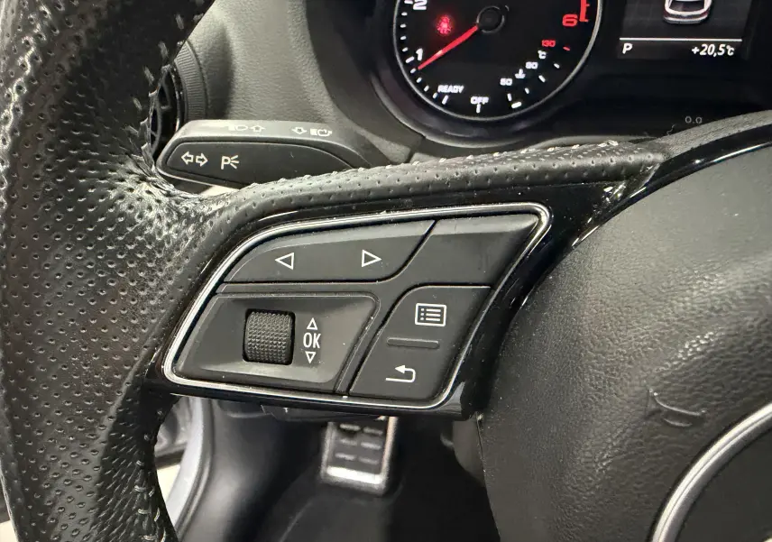 Gros plan sur les commandes multifonctions à gauche du volant perforé noir de l'Audi Q2 gris, avec compte-tours en arrière-plan.
