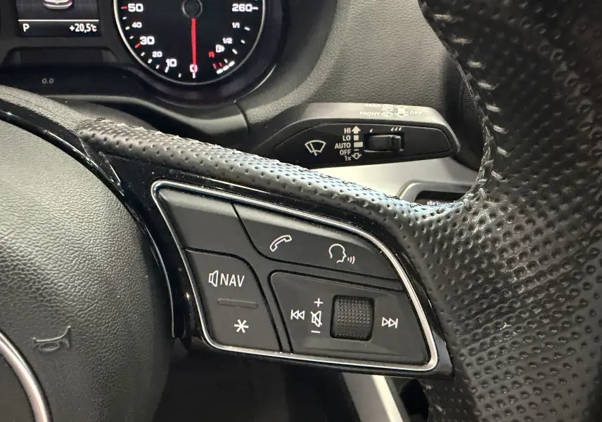 Gros plan sur les commandes multifonctions au volant en cuir perforé noir de l'Audi Q2 gris, avec tableau de bord partiellement visible.