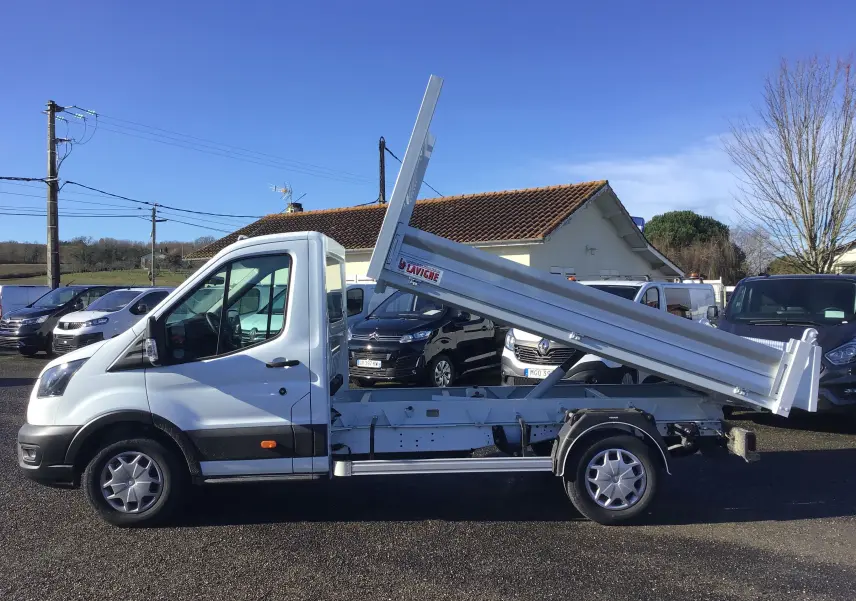Vue de profil côté gauche d'un Ford Transit blanc avec benne relevée sur un parking ensoleillé.