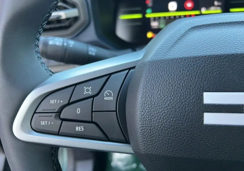 Gros plan sur les commandes au volant côté gauche du Dacia Duster 2025 vert cèdre, avec affichage numérique en arrière-plan.