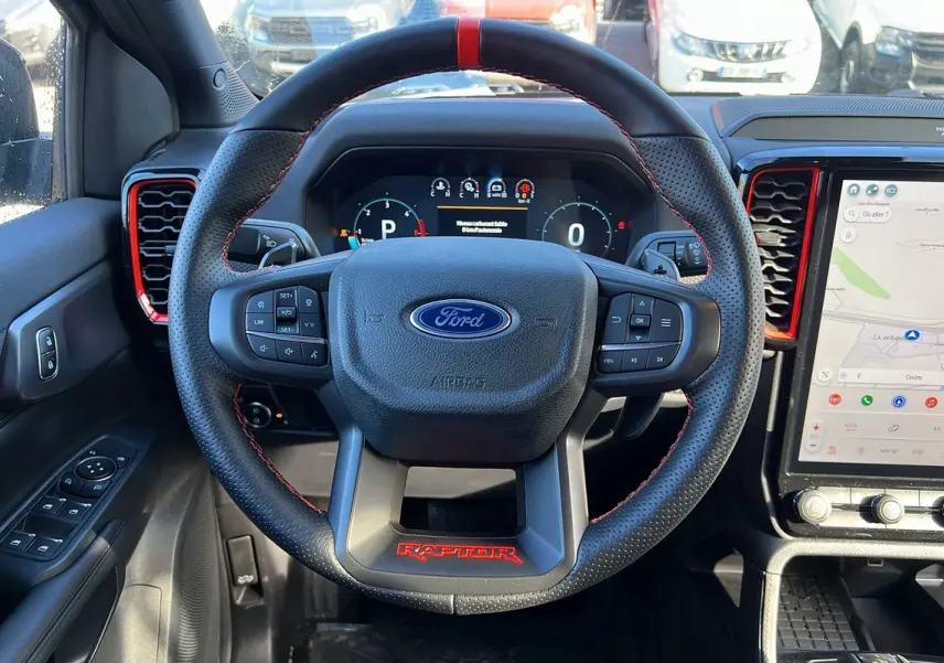 Vue intérieure centrée sur le volant noir perforé avec logo Ford et inscription rouge Raptor, tableau de bord digital et écran tactile.