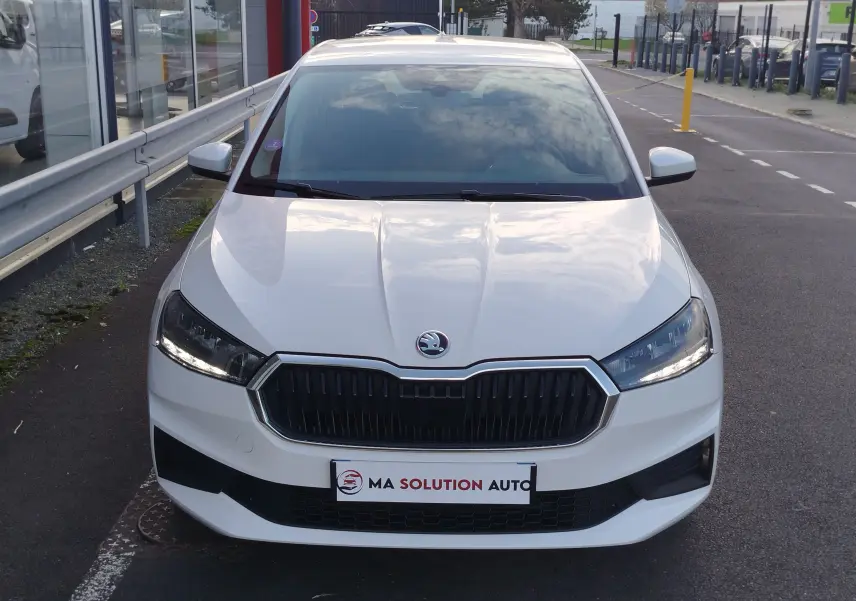 Vue avant d'une Skoda Fabia blanche Lune 2022 avec calandre noire et phares LED allumés sur parking urbain.