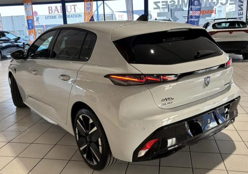 Vue 3/4 arrière droite d'une Peugeot 308 blanche hybride avec jantes bi-tons et bandeau noir brillant arrière en showroom.